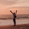 man in white shirt and black shorts standing on seashore during sunset