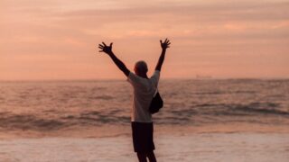 man in white shirt and black shorts standing on seashore during sunset